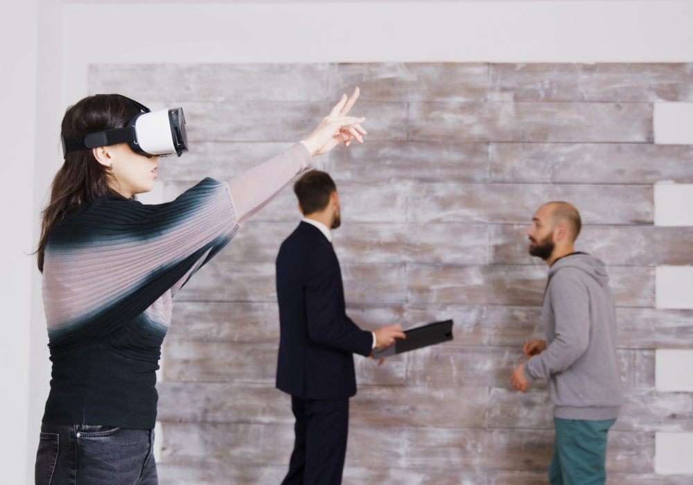 Female designer with virtual reality goggles in empty apartment while real estate agent is talking with the homeowner in the background.